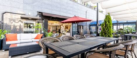 Un espace extérieur aménagé en terrasse, avec une table à manger, des chaises en osier, un canapé avec des coussins orange et un parasol rouge.