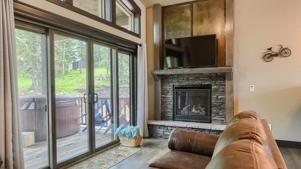 A living room with a fireplace, a large window, and a sliding glass door leading to an outdoor area with a hot tub. - New Venture- 3BR Cabin by Terry Peak (Lead)