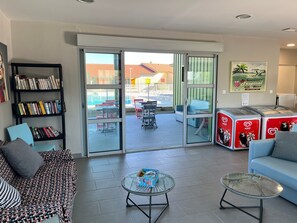 A modern living room with a sofa, a coffee table, a bookshelf, and a sliding glass door leading to an outdoor seating area. - Appartement proche plage | Piscine + Sauna (Marseillan-Plage)