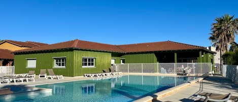 Ein Schwimmbecken mit Liegestühlen, ein grünes Gebäude mit Fenstern und ein klarer, blauer Himmel.