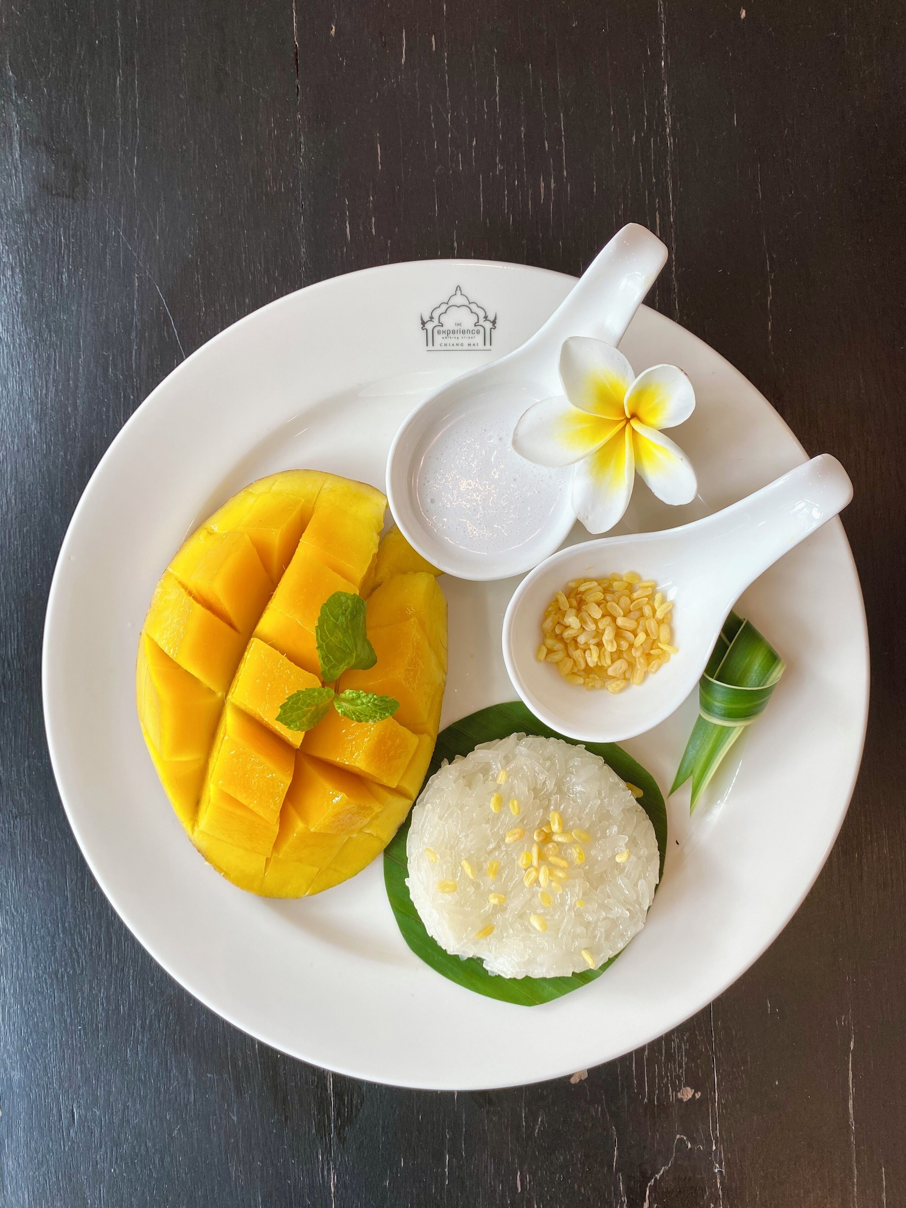 Une assiette avec des tranches de mangue, un bol de riz garni de haricots mungo, une cuillère blanche ornée d’une fleur et une feuille verte.