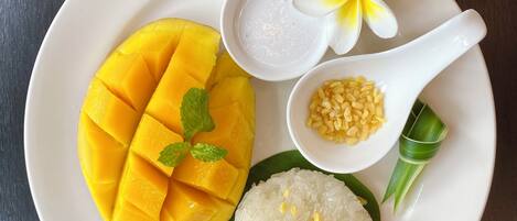 Un plato con mango en rodajas, un tazón de arroz cubierto de frijoles mung, una cuchara blanca con una flor, y una hoja verde.