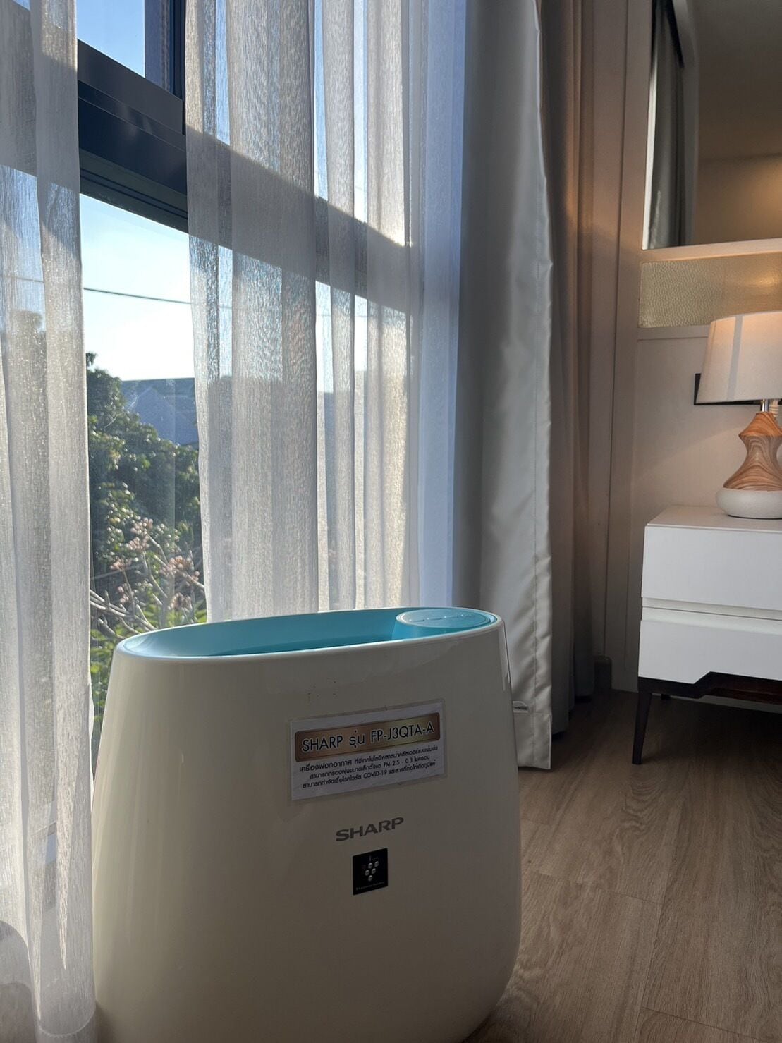 Una habitación con un purificador de aire Sharp, una mesita de noche blanca con una lámpara y una ventana con cortinas translúcidas.