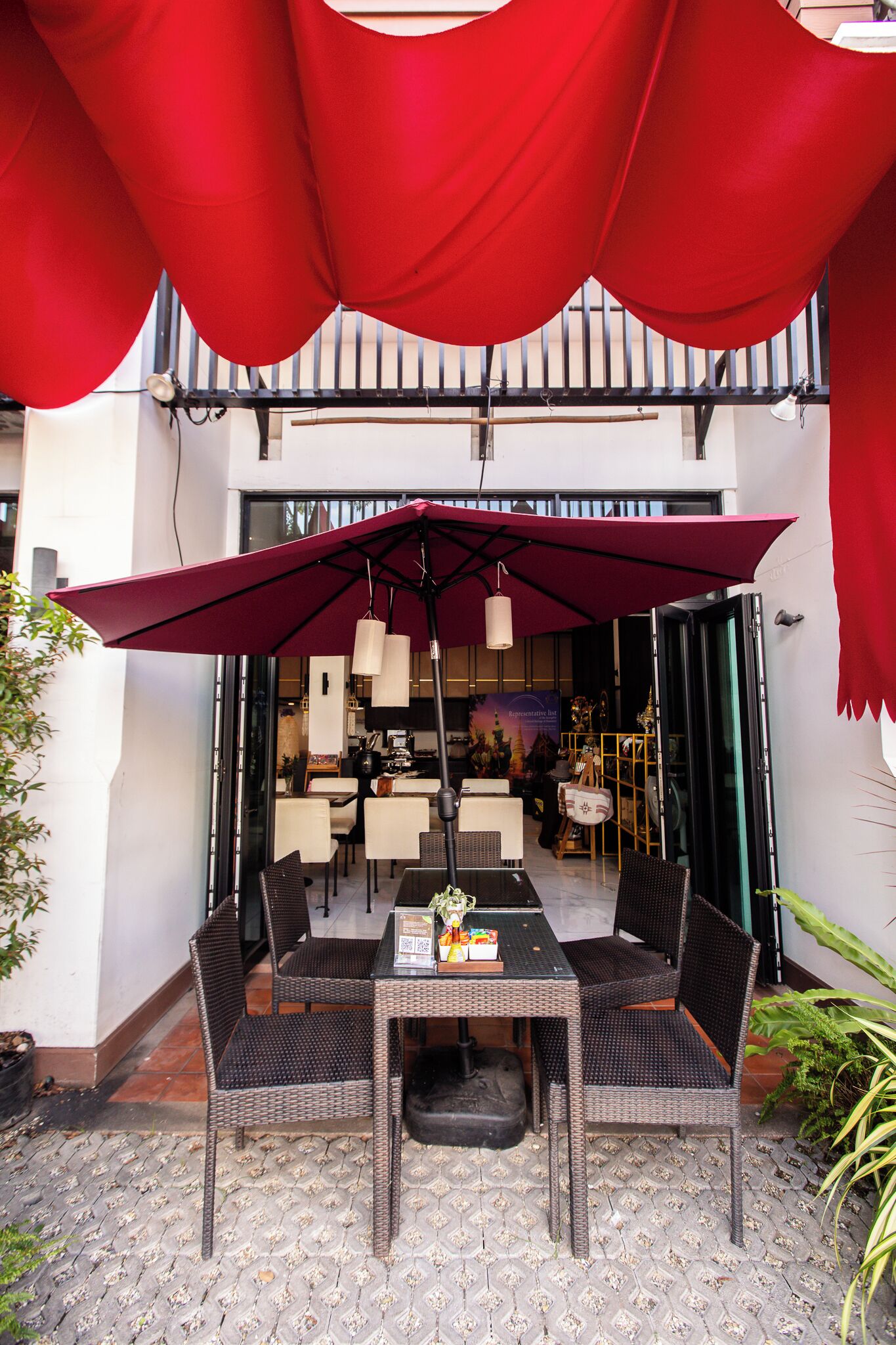 An outdoor dining area with wicker tables and chairs, a large red umbrella, and a view into the restaurant interior.