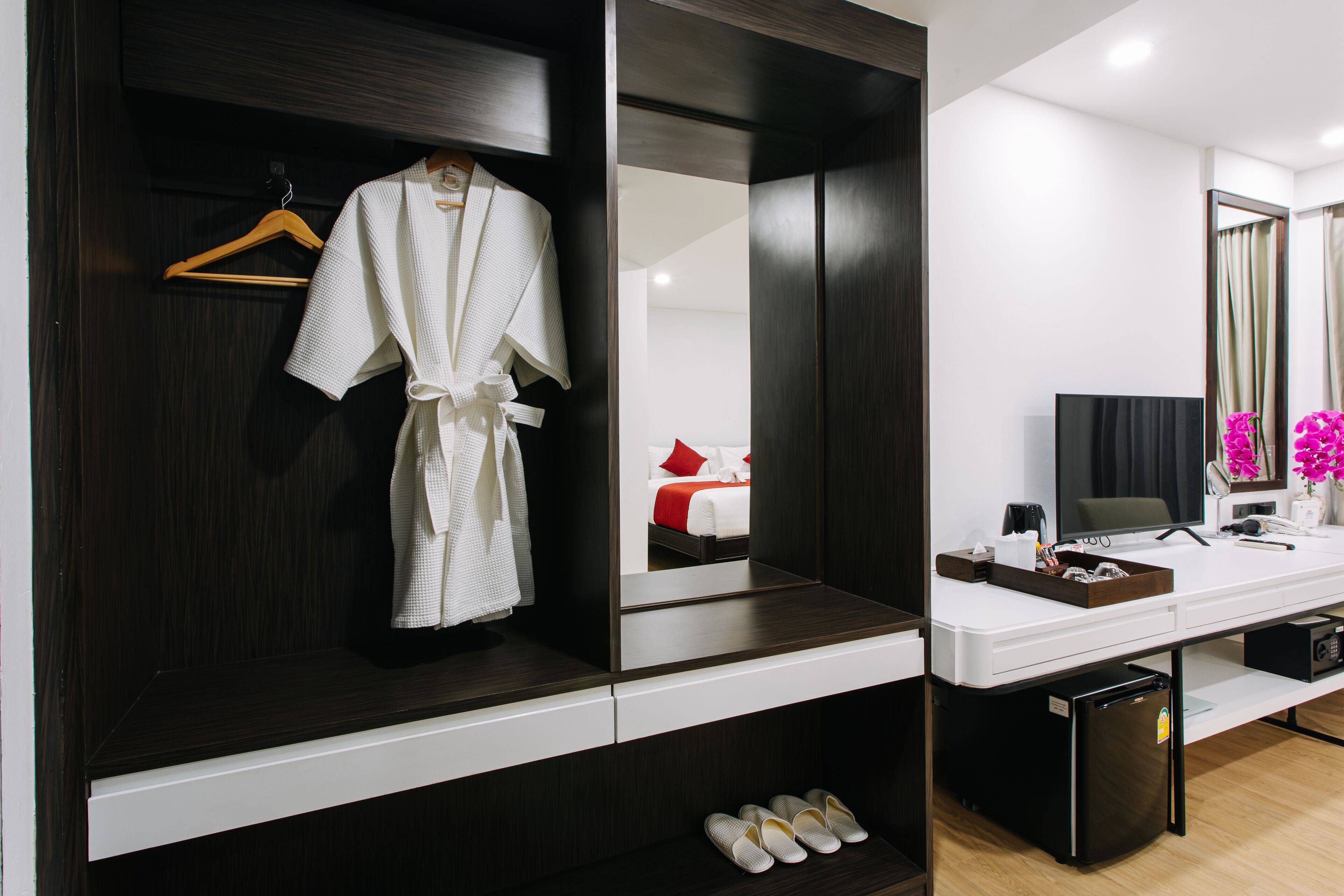 Ein Hotelzimmer mit einem dunklen Holzschrank, einem weißen Schreibtisch mit einem Computer, einem kleinen Kühlschrank und einem Spiegel, der ein Bett mit roten Kissen reflektiert.