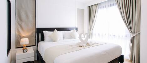 A neatly made bed with a heart-shaped towel arrangement, a bedside table with a lamp, and a large window with curtains.