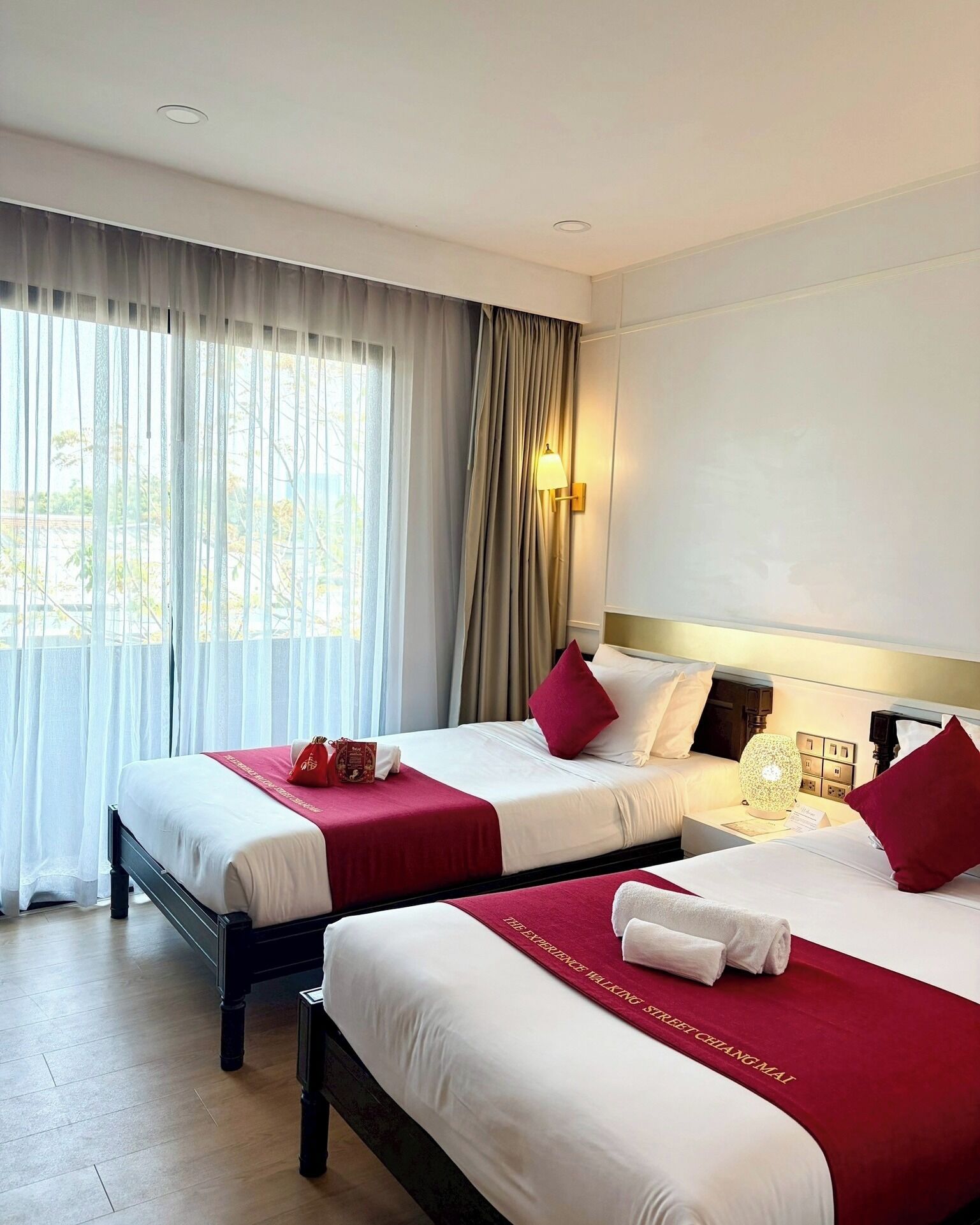 Habitación de hotel con dos camas, un ventanal con cortinas translúcidas, una lámpara de pared y una mesita de noche con lámpara.