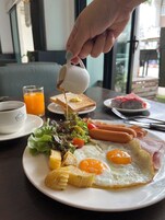 A breakfast plate with eggs, sausages, bacon, and toast, with a hand pouring syrup.