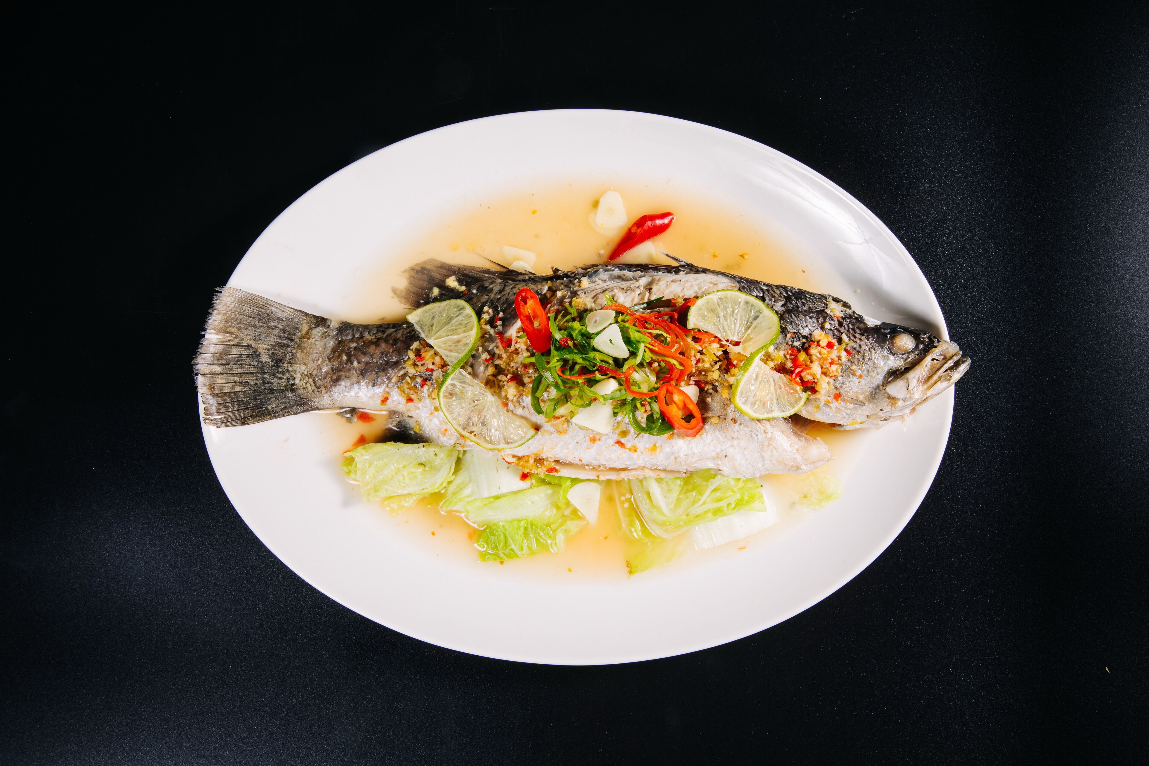 Une assiette blanche sur laquelle est présenté un poisson entier, agrémenté de citron vert, de piment et d’herbes, et servi sur une table noire.