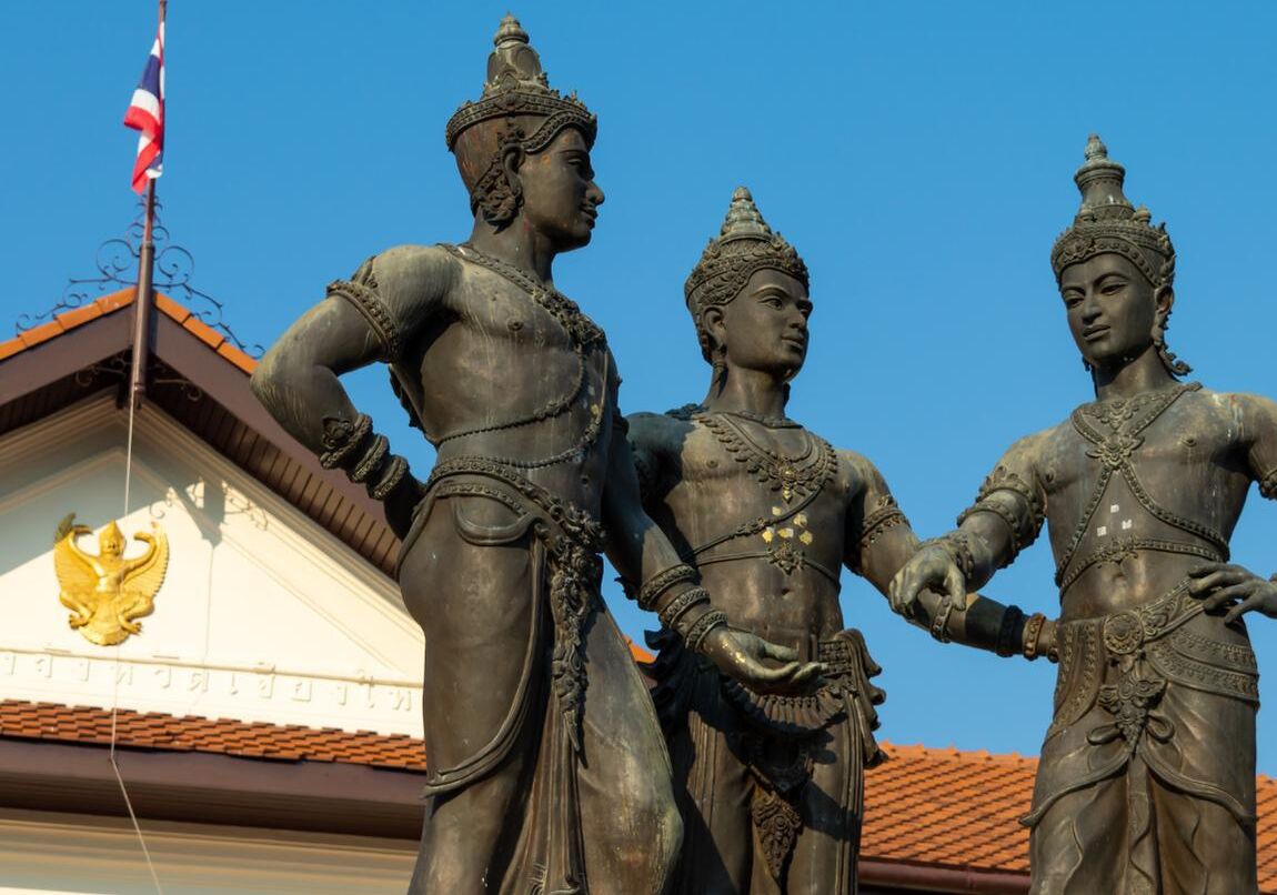 A statue of three figures in traditional attire, with a building featuring a golden emblem in the background.
