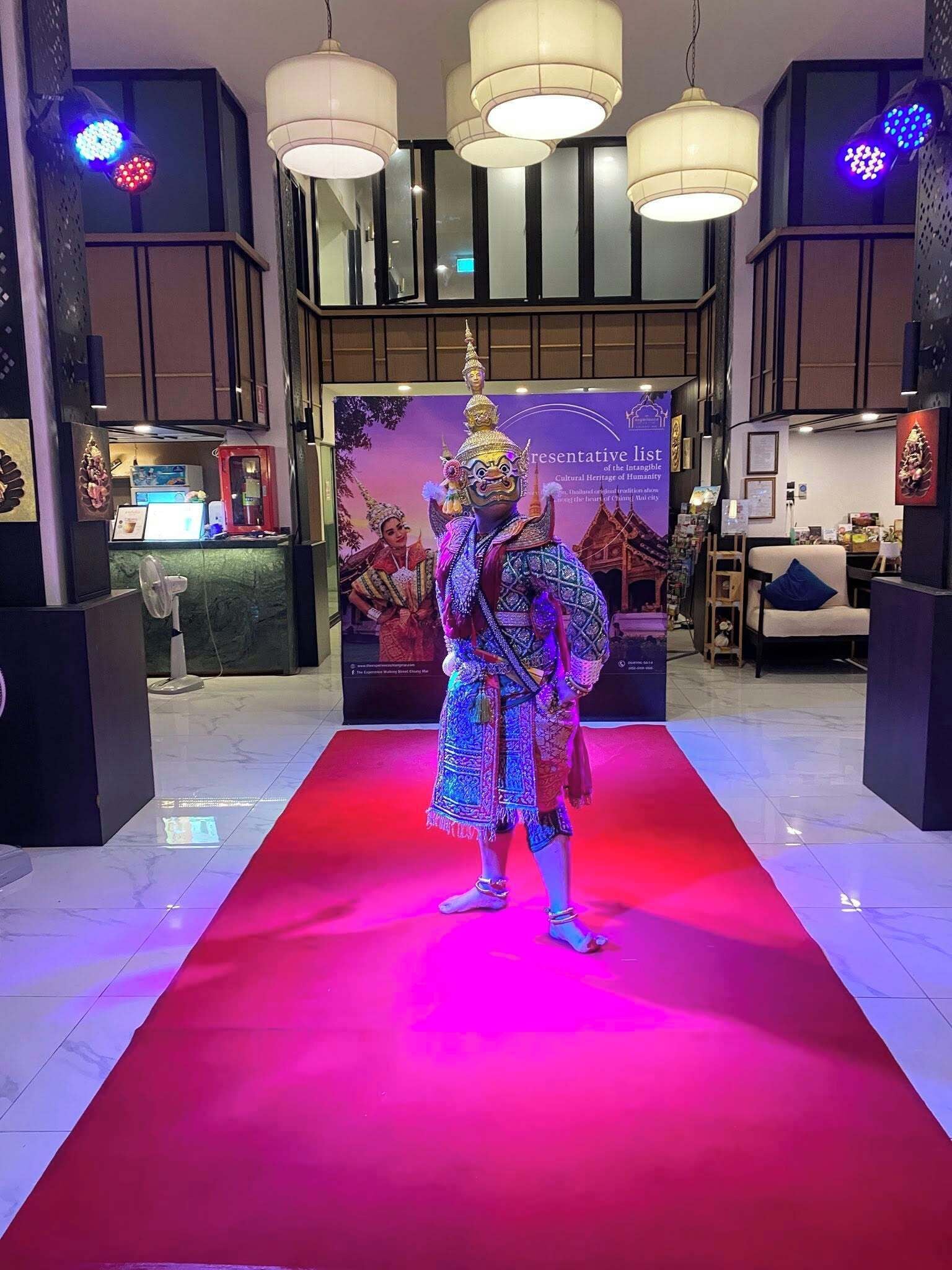 Una persona vestida con traje tradicional sobre una alfombra roja en un vestíbulo con iluminación moderna y un fondo que muestra información sobre el patrimonio cultural.