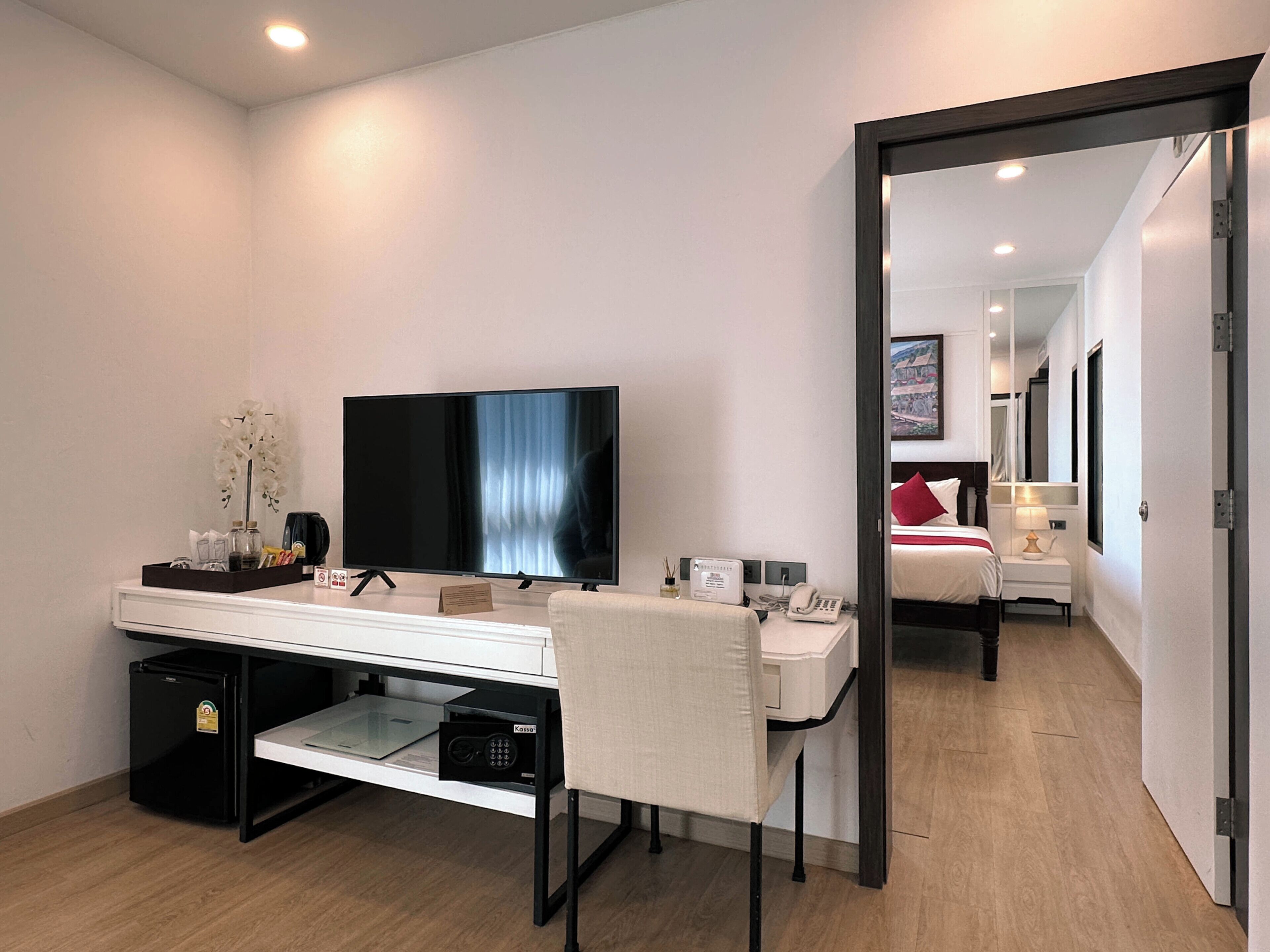 A modern hotel room with a flat-screen TV, a desk with a computer, a small fridge, a bedside table with a lamp, and a bed with a red pillow.