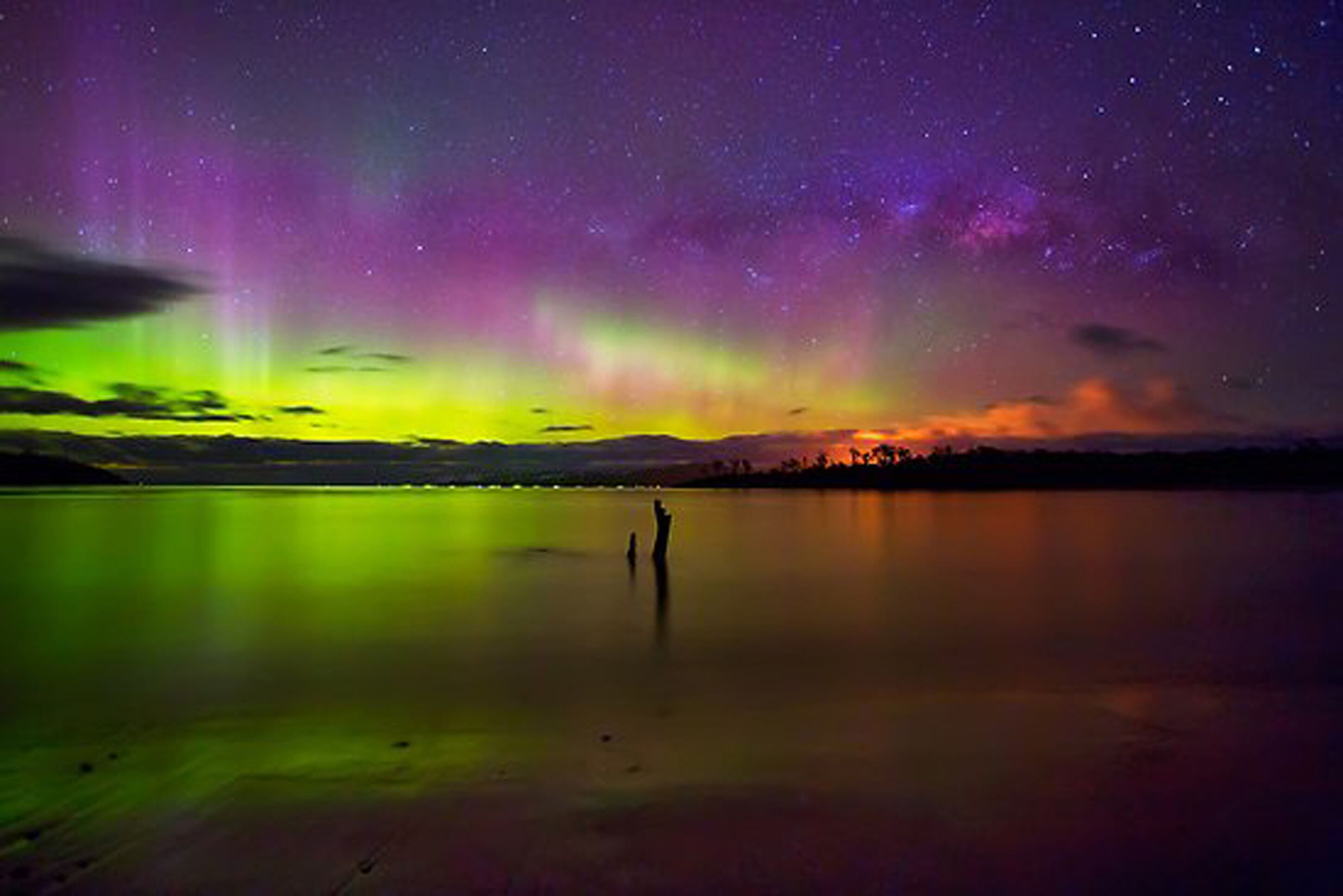 A nighttime view of a calm lake with a person standing in the center, reflecting the vibrant colors of the aurora borealis in the sky.