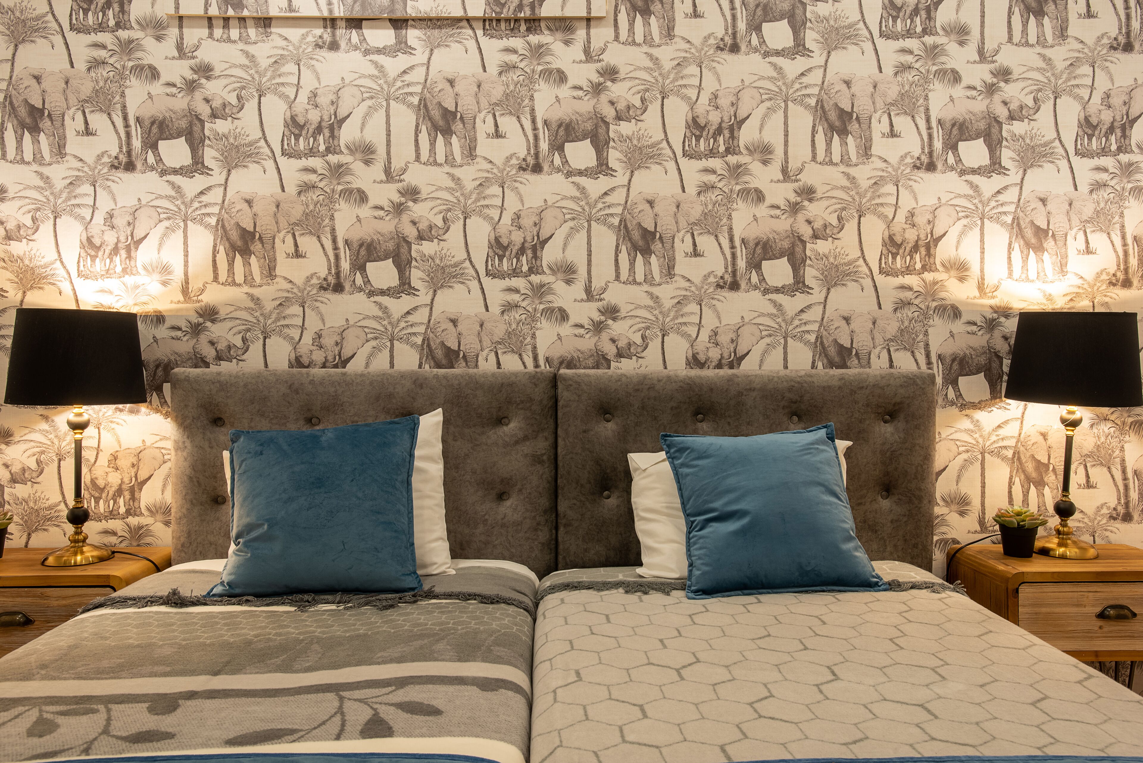 Une chambre à coucher avec une tête de lit capitonnée, deux coussins bleus et blancs, une couvre-lit à motifs et un mur recouvert d’un papier peint sur le thème de la faune.