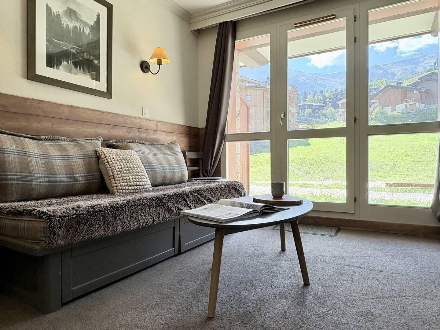 Een kamer met een bank, een tafel, een lamp en uitzicht op een berglandschap door een groot raam.