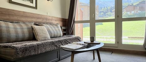 A room with a sofa, a table, a lamp, and a view of a mountain landscape through a large window.