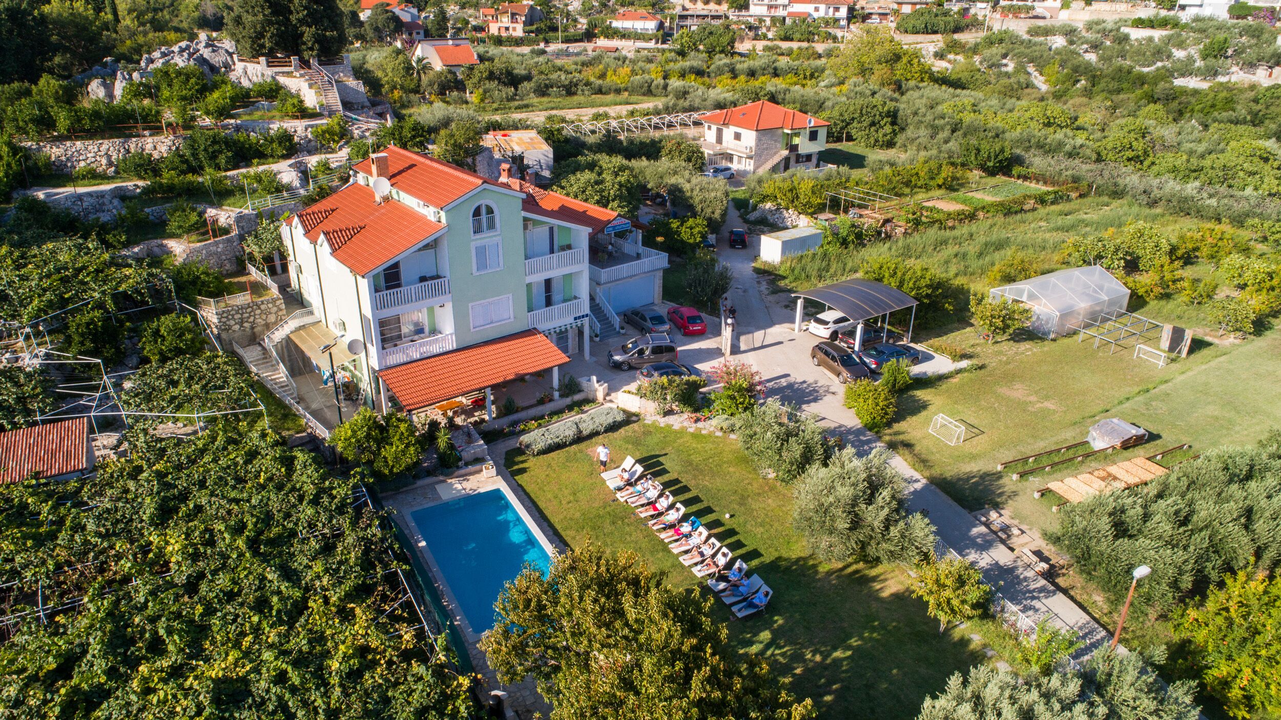 An aerial view of a residential area with a large house featuring a red-tiled roof, a swimming pool, and a garden with sun loungers.