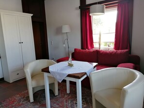 A room with a white table, two white chairs, a red sofa, and a window with red curtains.