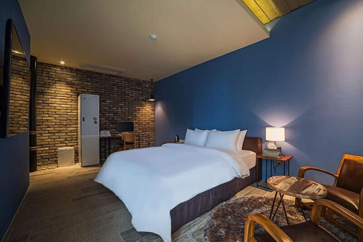 A hotel room with a large bed, a desk with a chair, a small table with a lamp, a brick wall, and a blue accent wall.