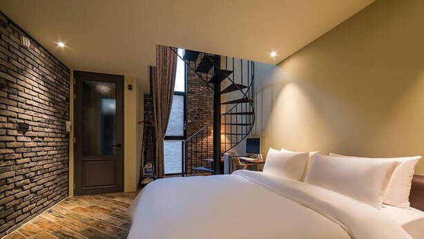 A modern hotel room with a large bed, a wooden floor, a brick accent wall, a staircase, and a desk with a chair. - Hotel March (Daejeon)