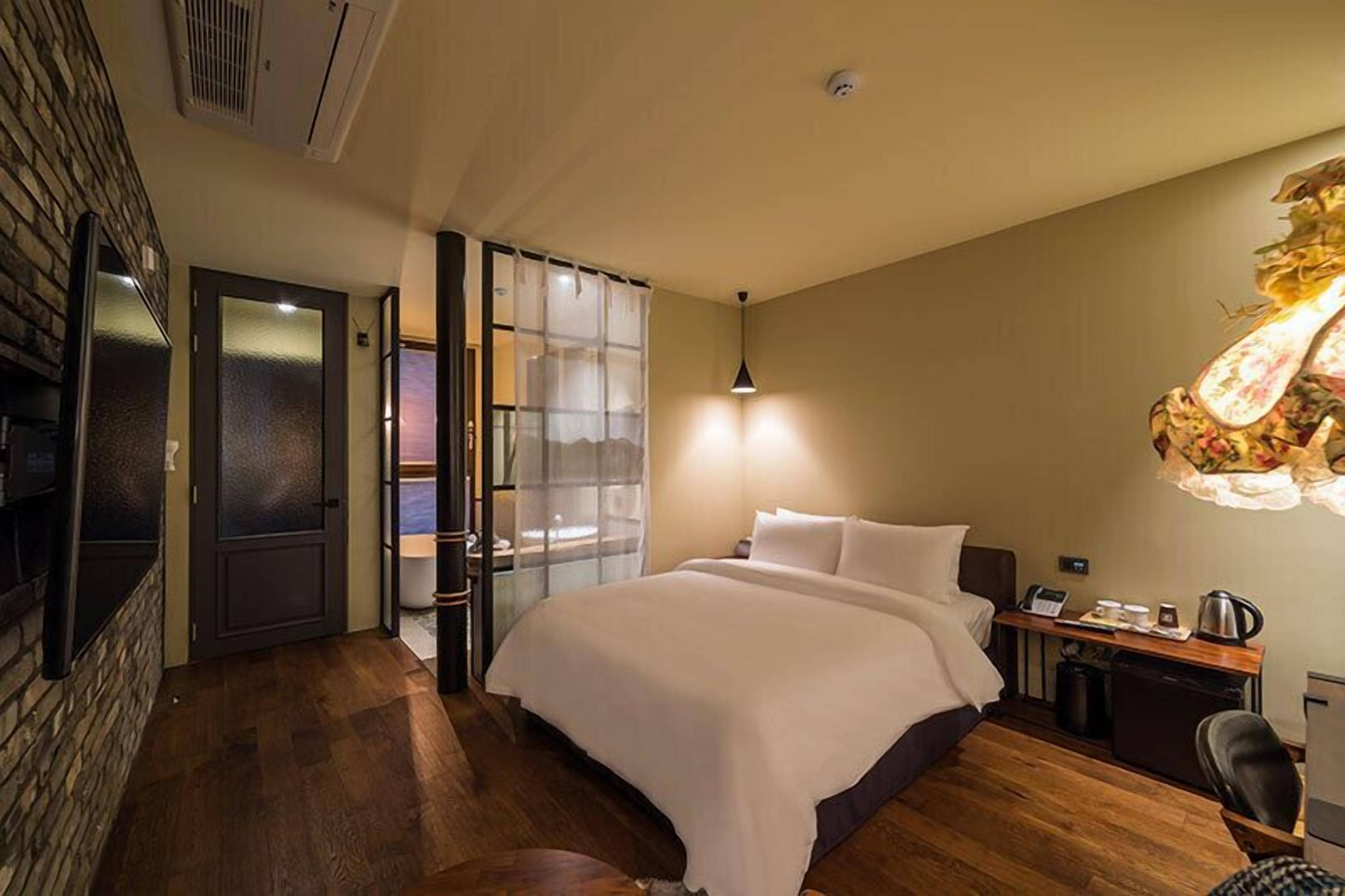 Habitación de hotel con cama, escritorio con lámpara, silla y una mesita con hervidor.