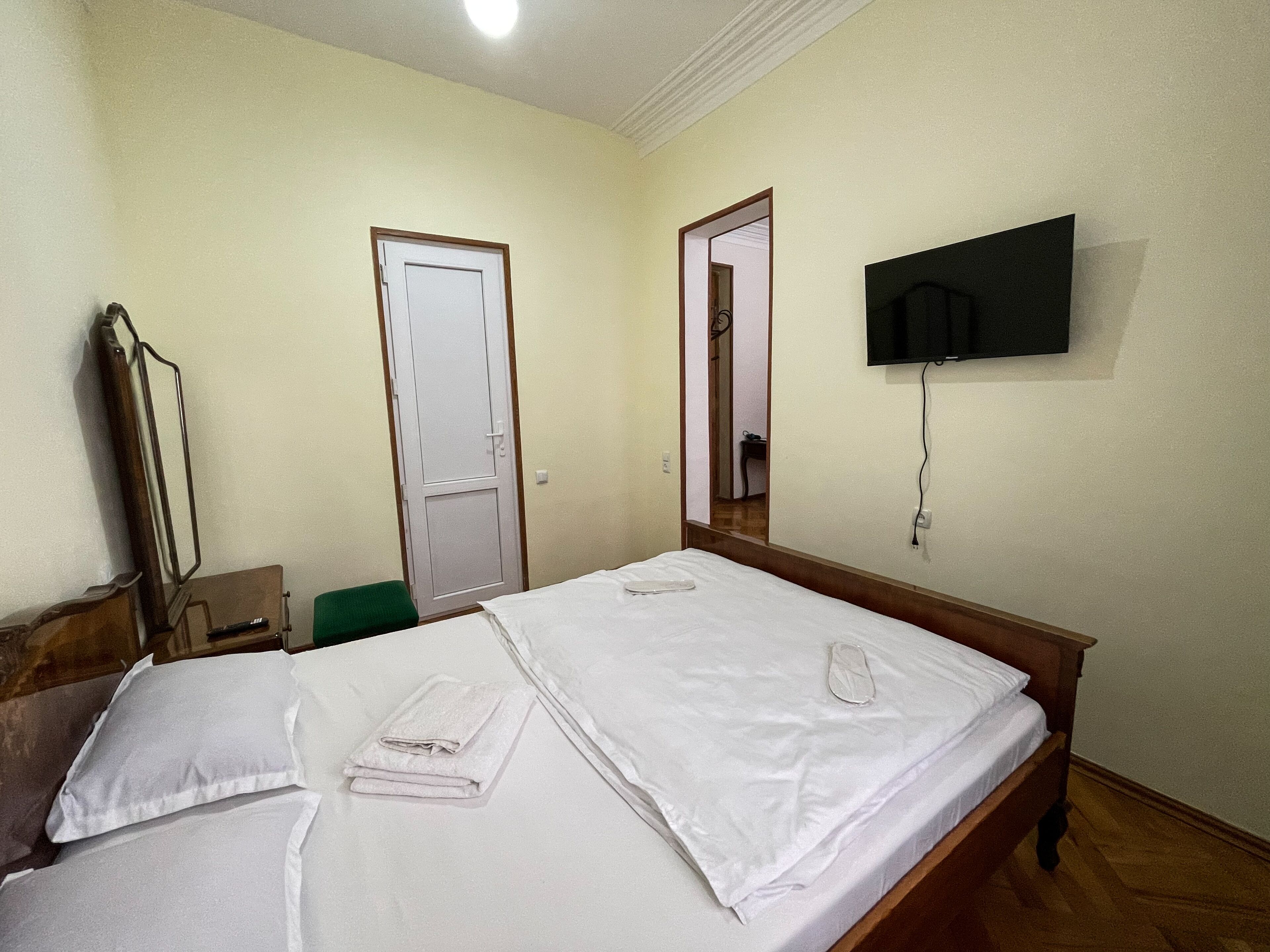 Un lit bien fait, avec du linge de lit blanc, un téléviseur à écran plat fixé au mur et une porte donnant sur une autre pièce.