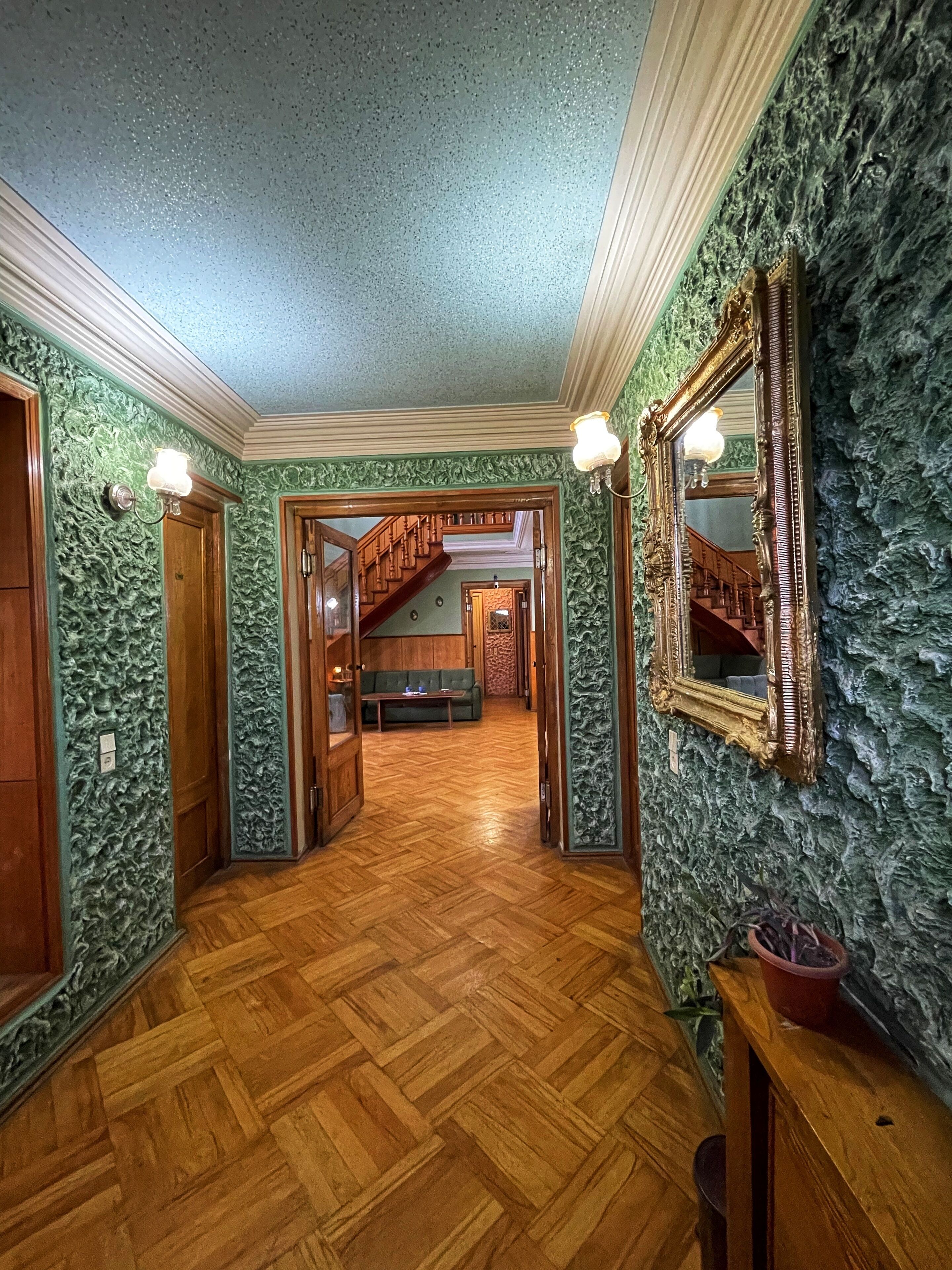A hallway with a wooden floor, green textured walls, and a mirror on the right.