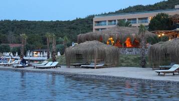 Een hotel aan het strand met rieten parasols, ligbedden en een zwembad.