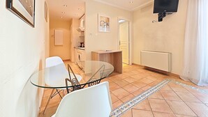 A modern living room with a glass dining table, white chairs, a TV mounted on the wall, and a kitchen area in the background. - Studio Commandant André 22 by Viva Riviera (Cannes)