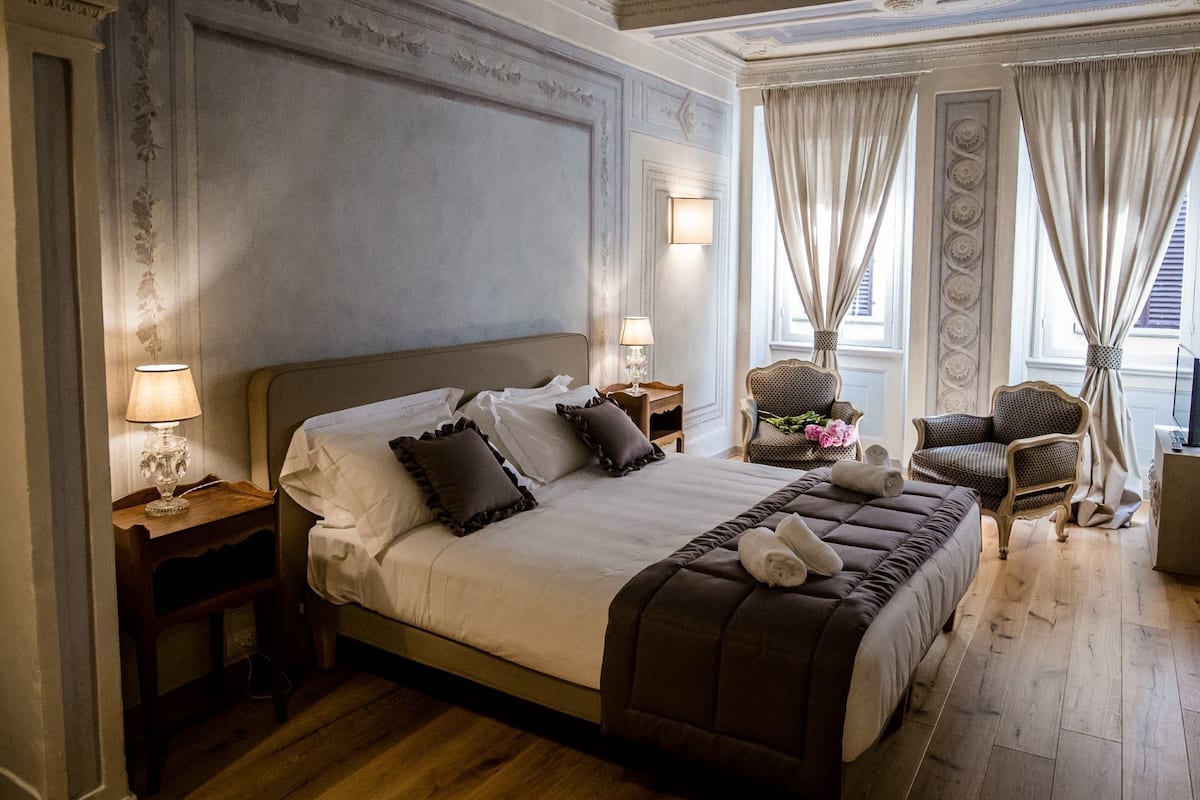 A spacious bedroom with a large bed, two nightstands, a sitting area with chairs, and a view of the city through large windows.