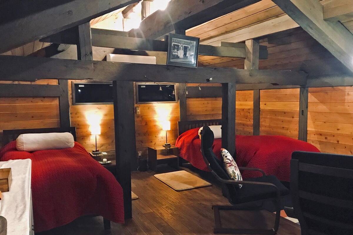 Una cabaña con dos camas, cada una con un edredón rojo, piso de madera y una mesita con lámpara.