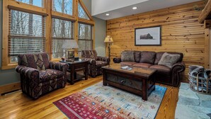 A cozy living room with a large window, wooden walls, a leather sofa, patterned armchairs, and a wooden coffee table.