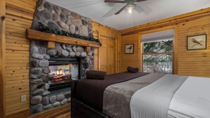 A cozy log cabin bedroom with a stone fireplace, wooden walls, a bed, and a window with a view of trees.