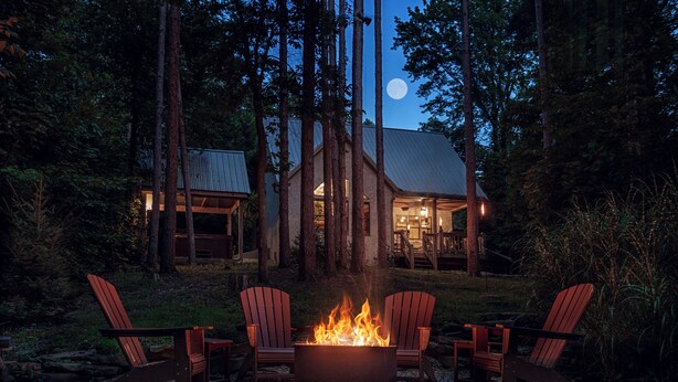 A cabin with a fire pit and four Adirondack chairs in front.