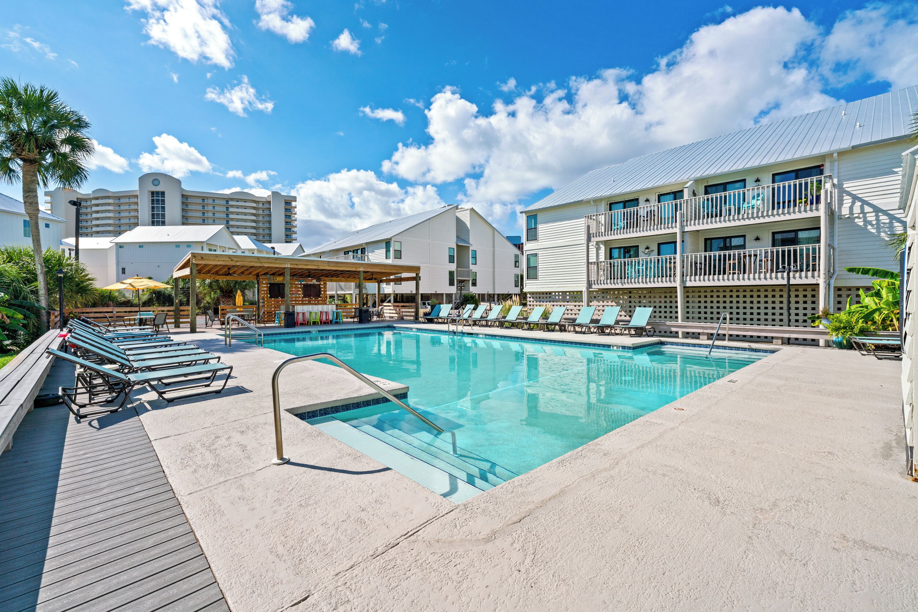 A swimming pool with lounge chairs, a covered seating area, and a building with multiple balconies in the background.
