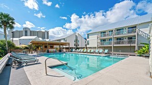 A swimming pool with lounge chairs, a covered seating area, and a building with multiple balconies in the background.