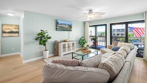 A spacious living room with a large sofa, a coffee table, and a ceiling fan. There is a large window with a view of the outside.