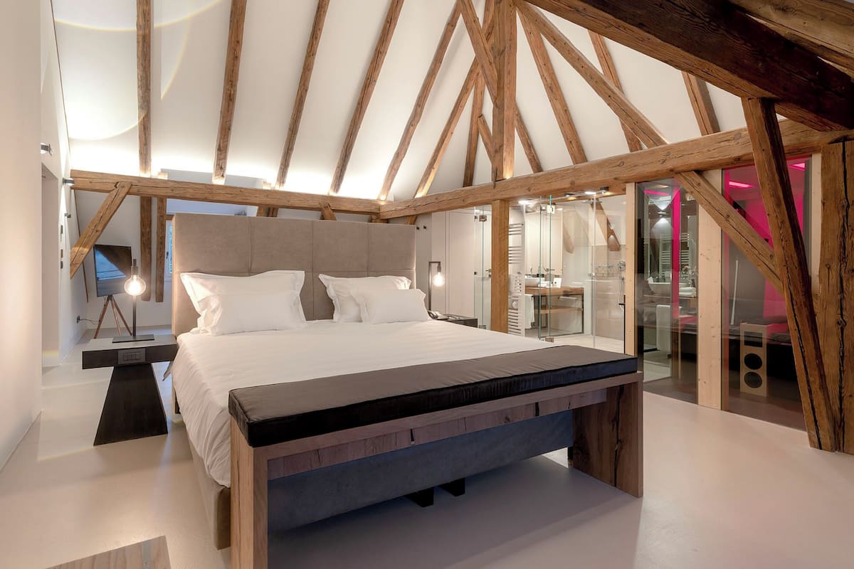Un dormitorio moderno con una cama grande, vigas de madera, un televisor y una zona de ducha con mampara de vidrio.