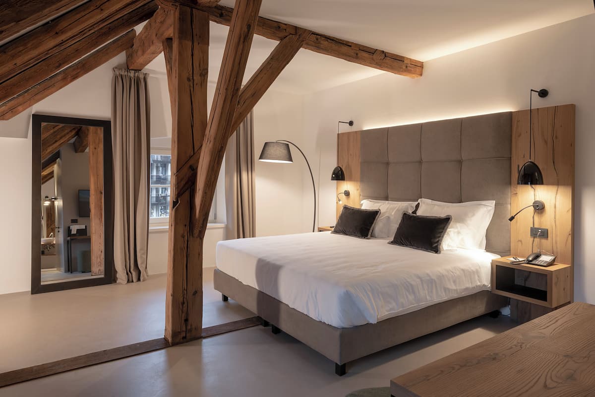 Un dormitorio moderno con una cama grande, vigas de madera, una mesita de noche con una lámpara y vistas a la ciudad a través de una ventana.