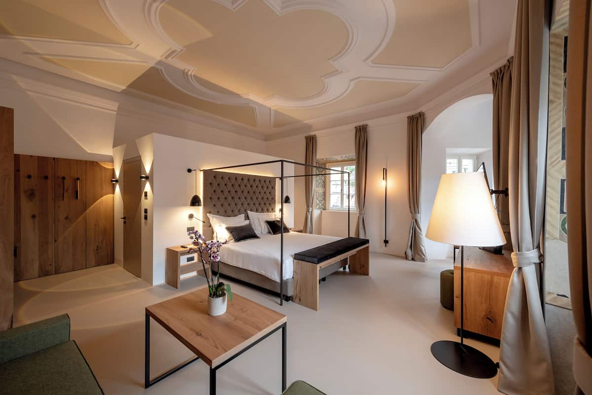 Un dormitorio moderno con cama con dosel, una mesita de noche con lámpara, un banco de madera y una pequeña mesa con una planta.