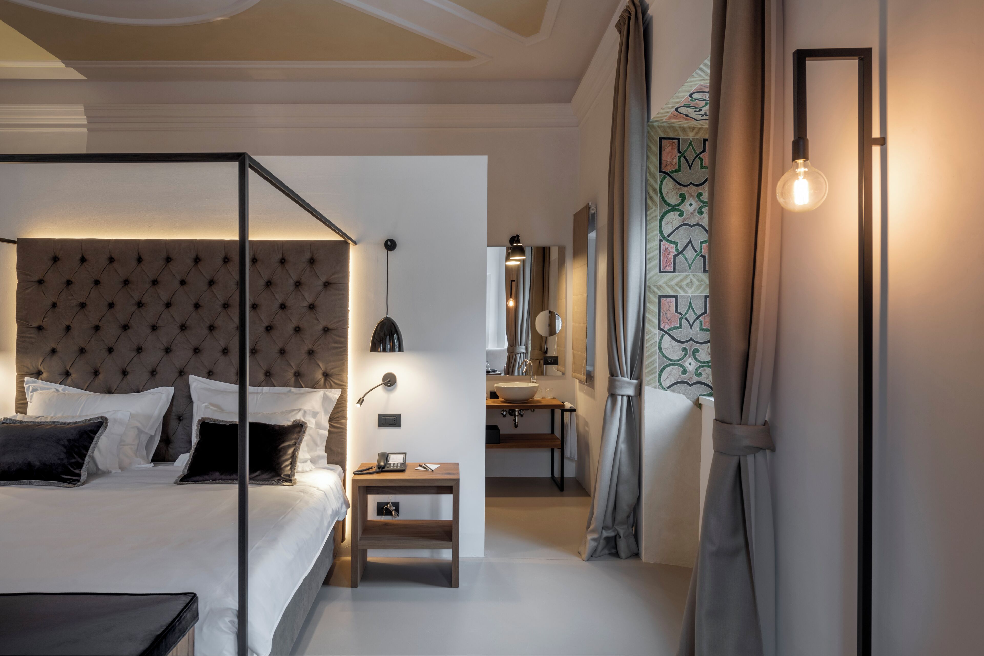 A modern bedroom with a tufted headboard, a bedside table with a lamp, and a view of a bathroom.