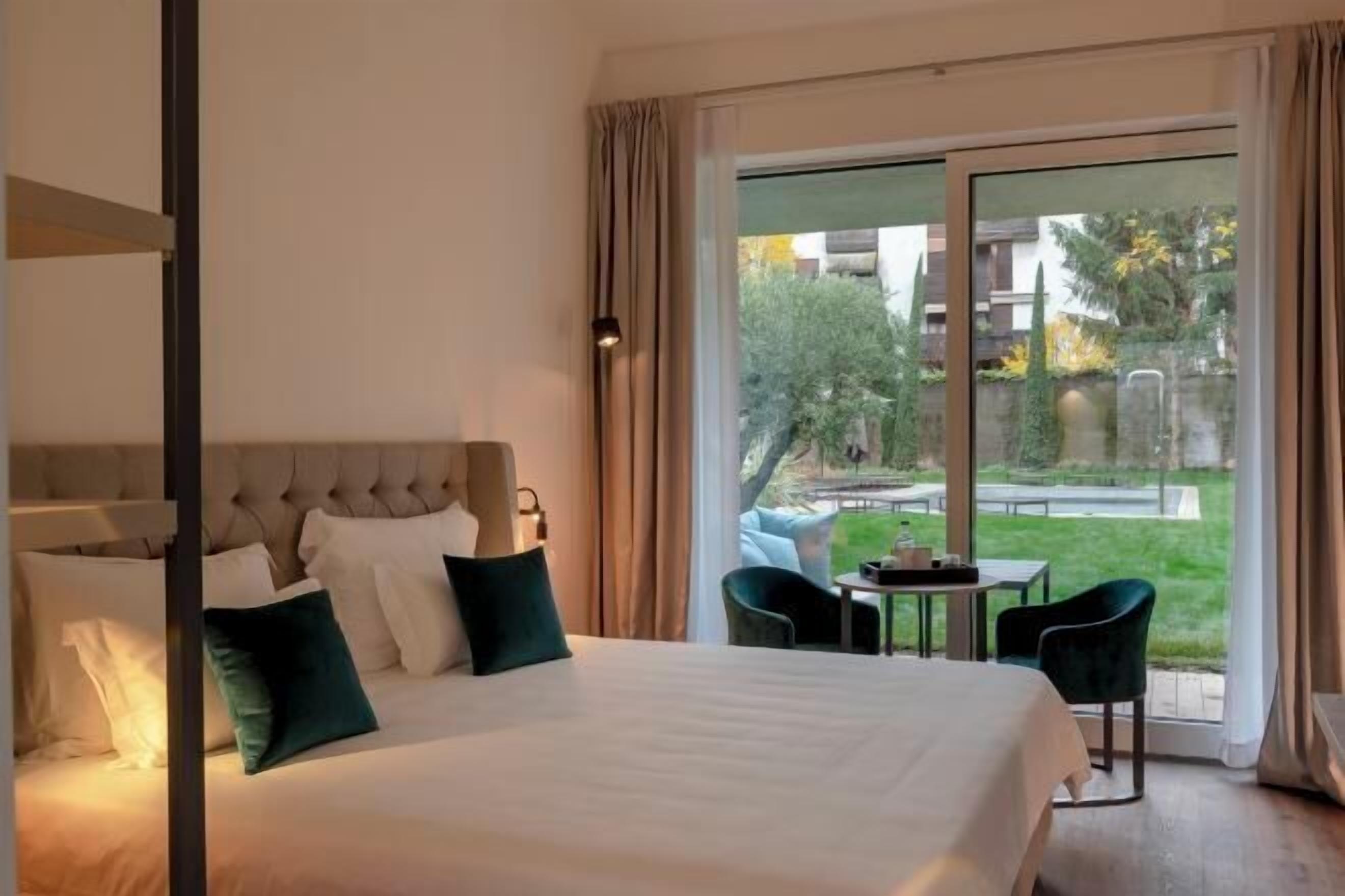 Une chambre d’hôtel avec un grand lit, une fenêtre donnant sur un jardin et une petite table avec des chaises.