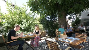 An outdoor café with wooden tables and benches, surrounded by greenery and buildings. - Pension Obstgarten (Achstetten)