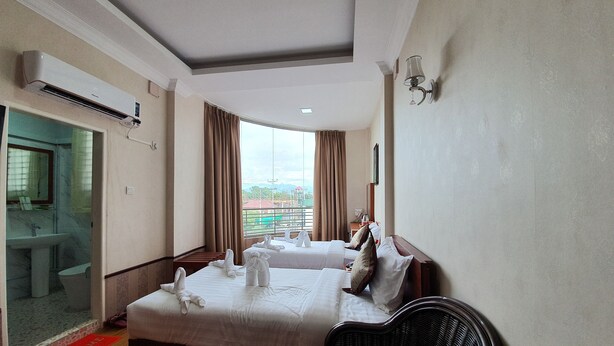 A hotel room with two beds, a bathroom, a view of a cityscape, and a red welcome mat. - Shwe Pe Ti Hotel (Mandalay)