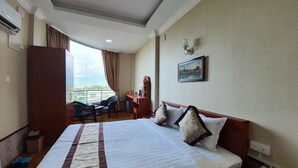 A hotel room with a large bed, a desk, a chair, and a view of a cityscape through the window. - Shwe Pe Ti Hotel (Mandalay)