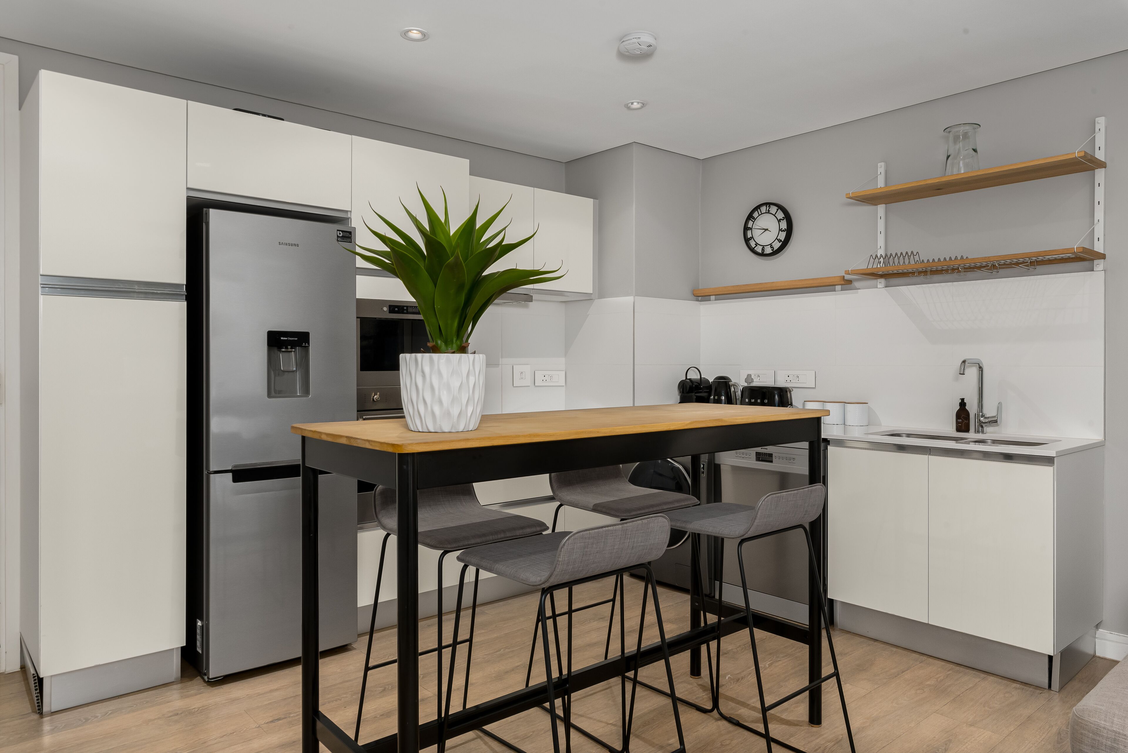 A modern kitchen with white cabinets, a stainless steel refrigerator, a wooden table with bar stools, a potted plant, a clock, and a coffee machine.