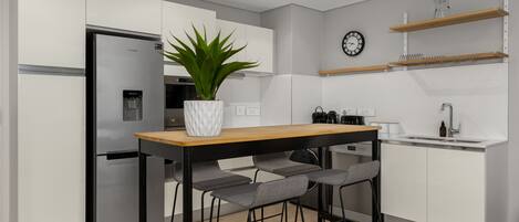 A modern kitchen with white cabinets, a stainless steel refrigerator, a wooden table with bar stools, a potted plant, a clock, and a coffee machine.