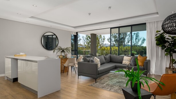 A modern living room with a grey sofa, a white desk, and a large window offering a view of trees. - Best of Both (Cape Town)