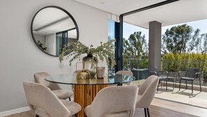 A modern dining area with a glass table, beige chairs, a round mirror, and a potted plant. - Best of Both (Cape Town)
