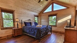 Un dormitorio con techo de madera, una cama con cubrecama azul, una mesita de noche, un aparador y dos ventanas.
